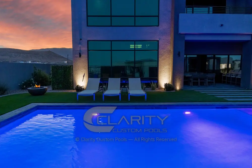 Pool lit violet-blue at dusk in front of a modern two-story home, with lounge chairs and a fire bowl on the deck