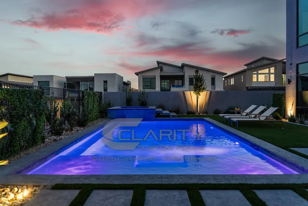Rectangular pool lit blue at dusk with a raised spa spilling into it, a tanning ledge and lounge chairs on the deck