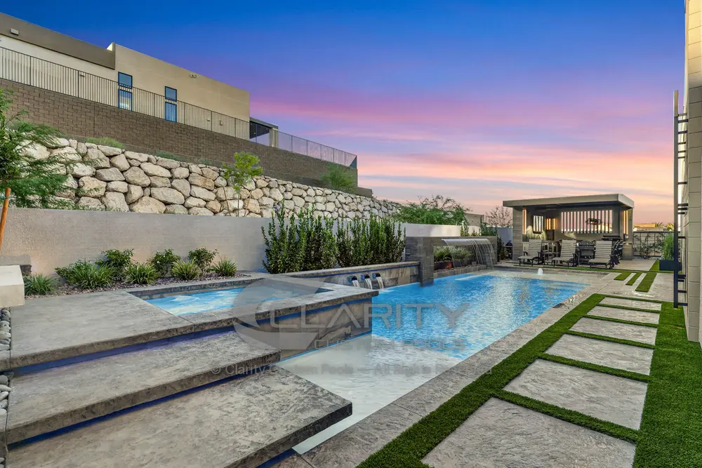 Long lap pool and raised spa at twilight with stepped concrete deck pads and a stacked-rock retaining wall