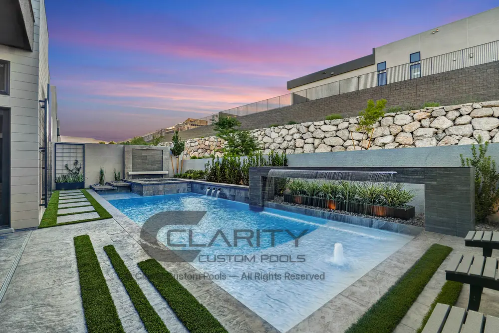 Pool at dusk with a rain-curtain water feature, a bubbler on the tanning ledge and a stacked-rock wall behind