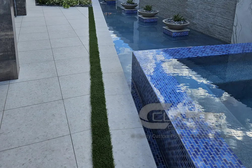 Raised spa clad in blue glass tile beside a long light-colored porcelain deck, with desert mountains beyond