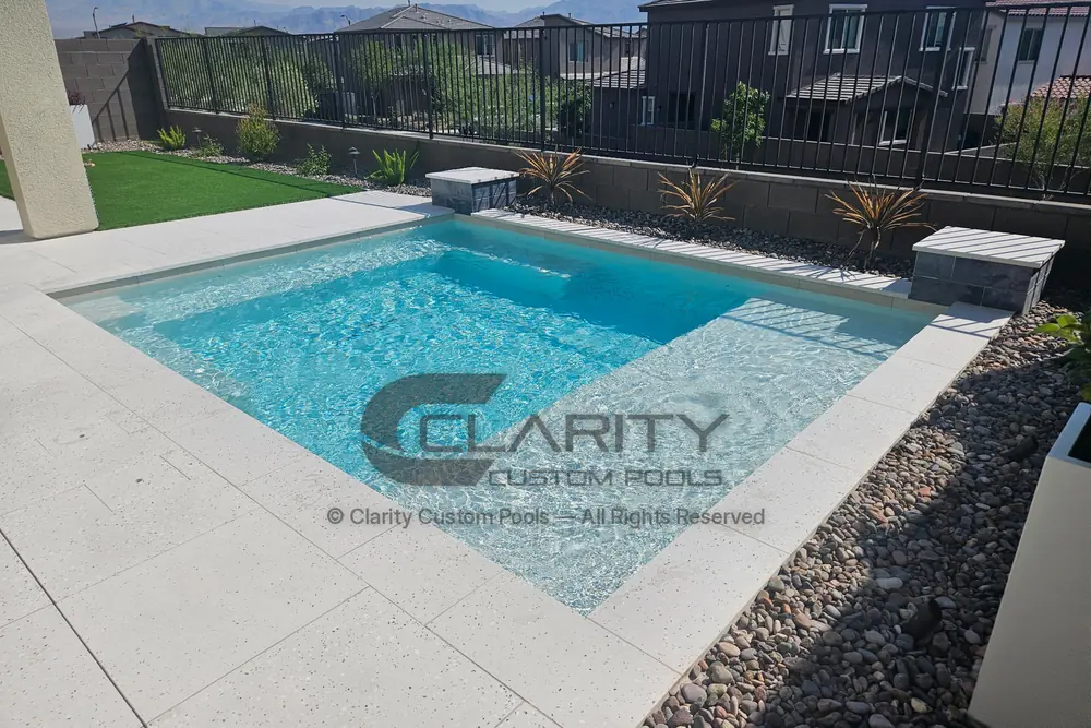 Compact rectangular pool with a wide shallow ledge, light deck pavers and a view fence along the back