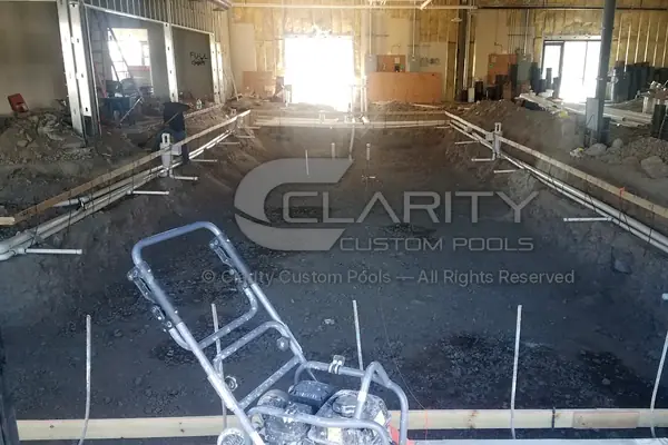 Excavated indoor commercial pool with wood forms along the edges and plumbing stub-ups rising from the floor