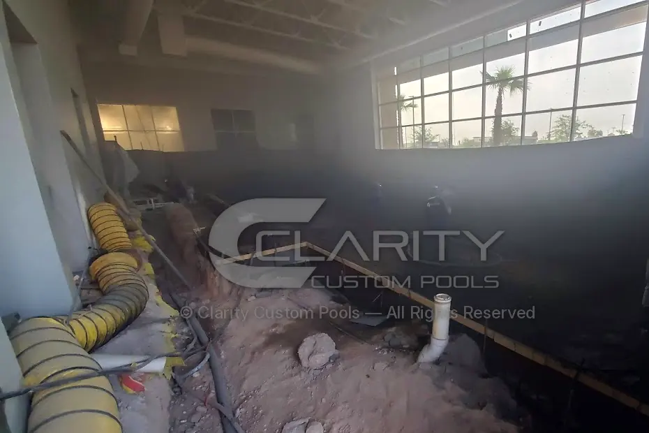 Dusty indoor pool shell during shotcrete application, with ventilation ducting along one side