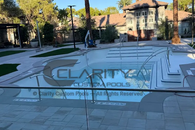 Finished outdoor community pool with an accessible pool lift, handrails and wide entry steps, surrounded by paver decking and a glass barrier